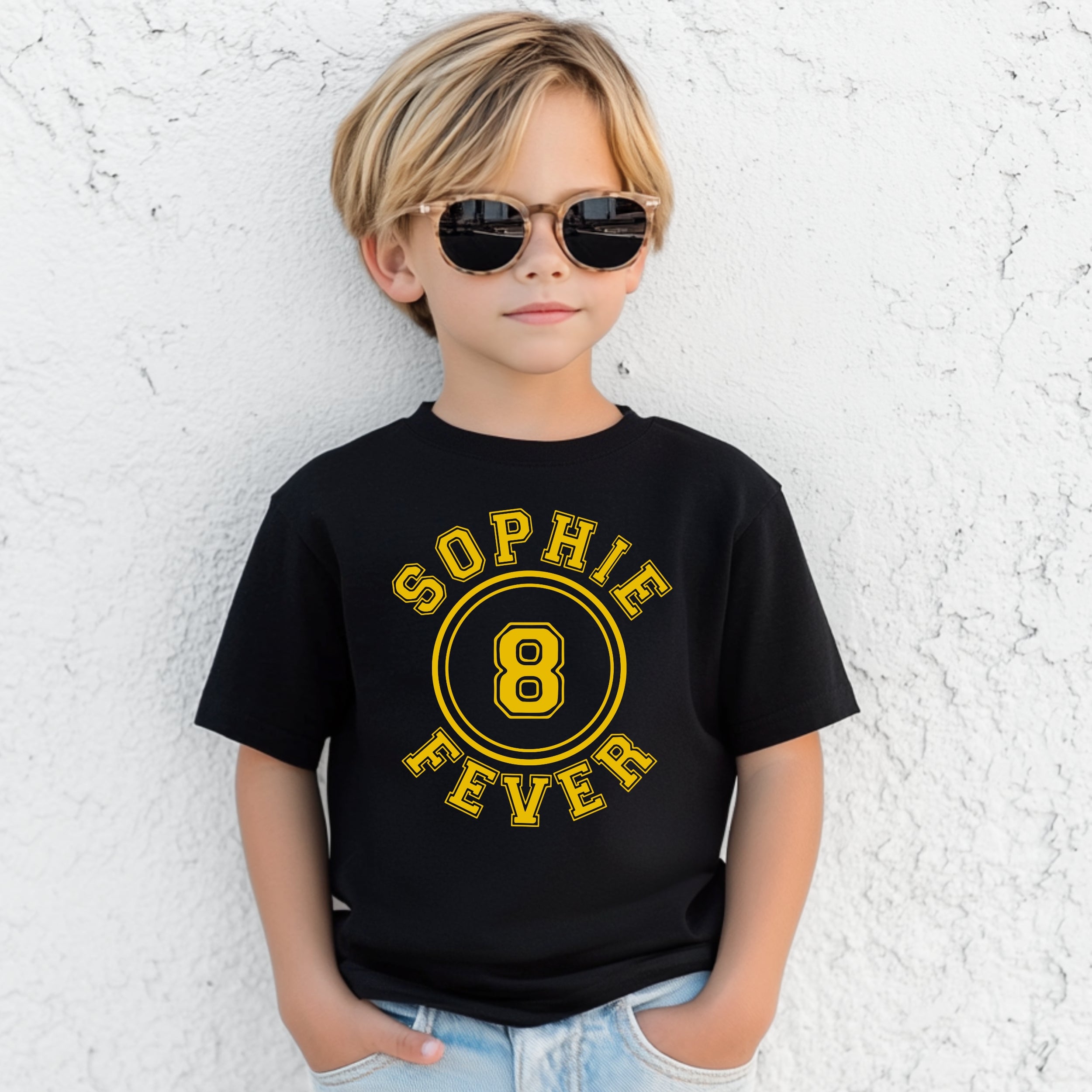 Child wearing a black t-shirt with 'Sophie 8 Fever' in yellow text against a textured white wall.