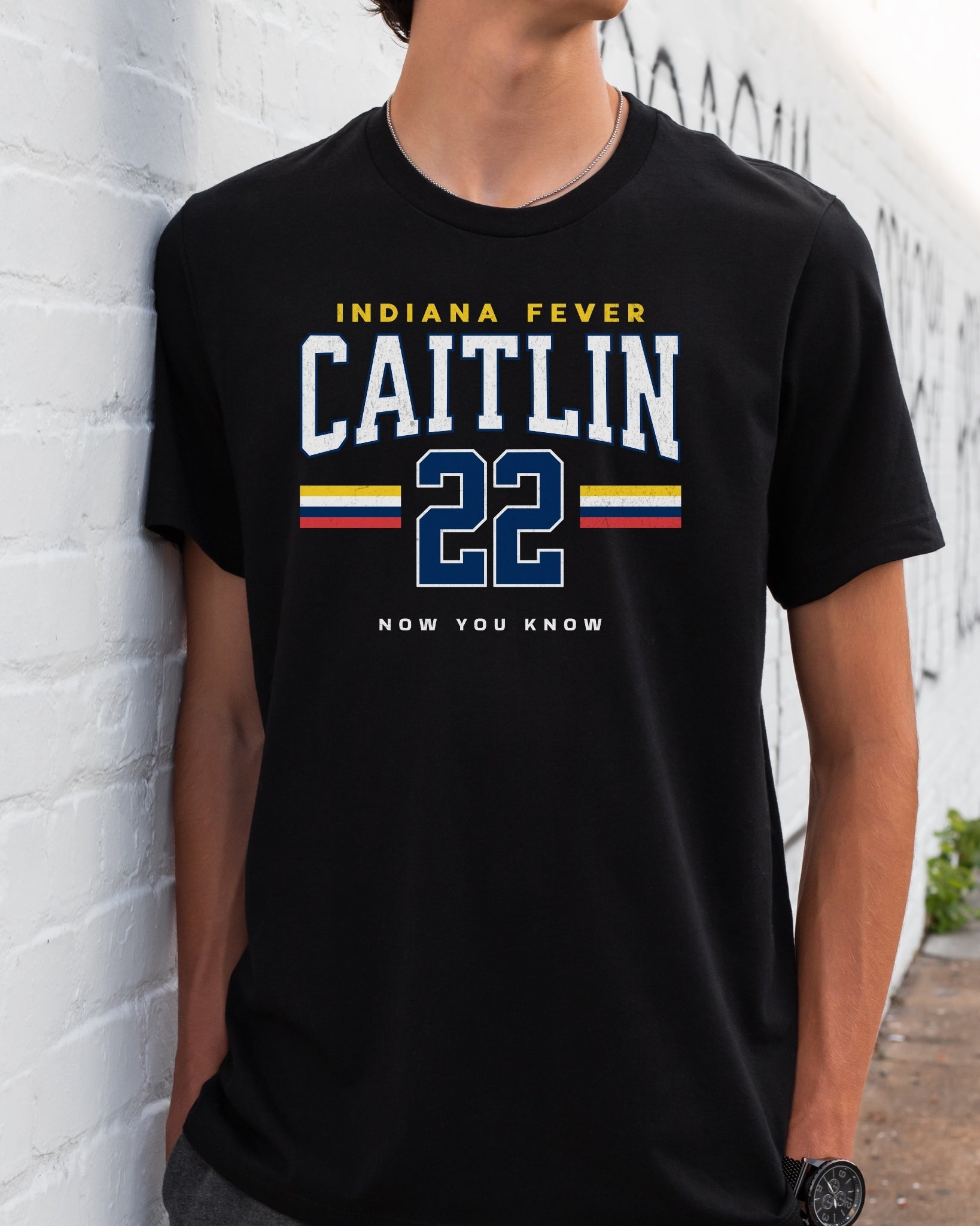 Person wearing a black t-shirt with 'Indiana Fever Caitlin 22 Now You Know' text against a white brick wall.