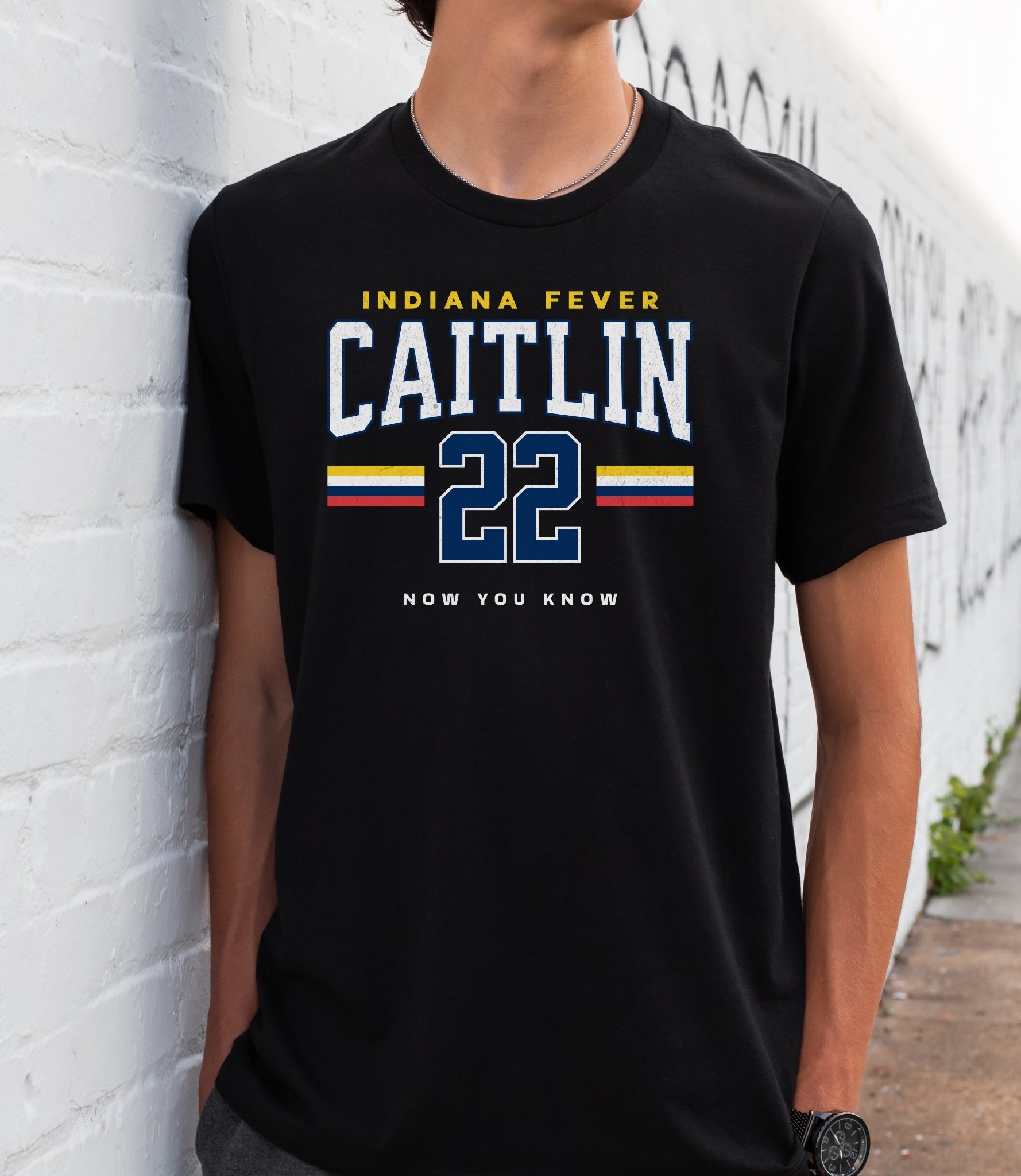 Person wearing a black t-shirt with 'Indiana Fever Caitlin 22 Now You Know' text against a white brick wall.