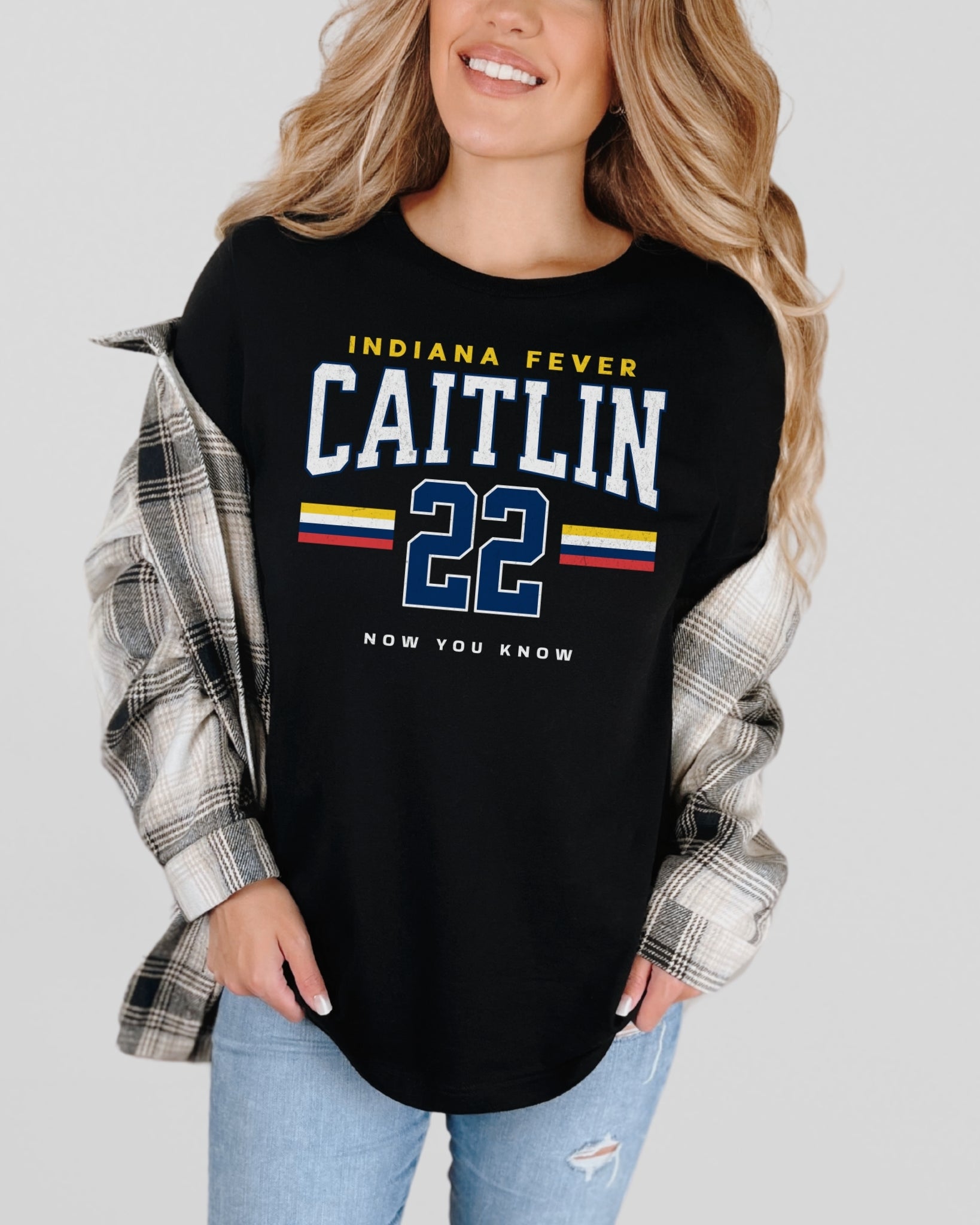 Woman wearing a black shirt with 'Indiana Fever Caitlin 22' text, flannel sleeves, and jeans on a light gray background.