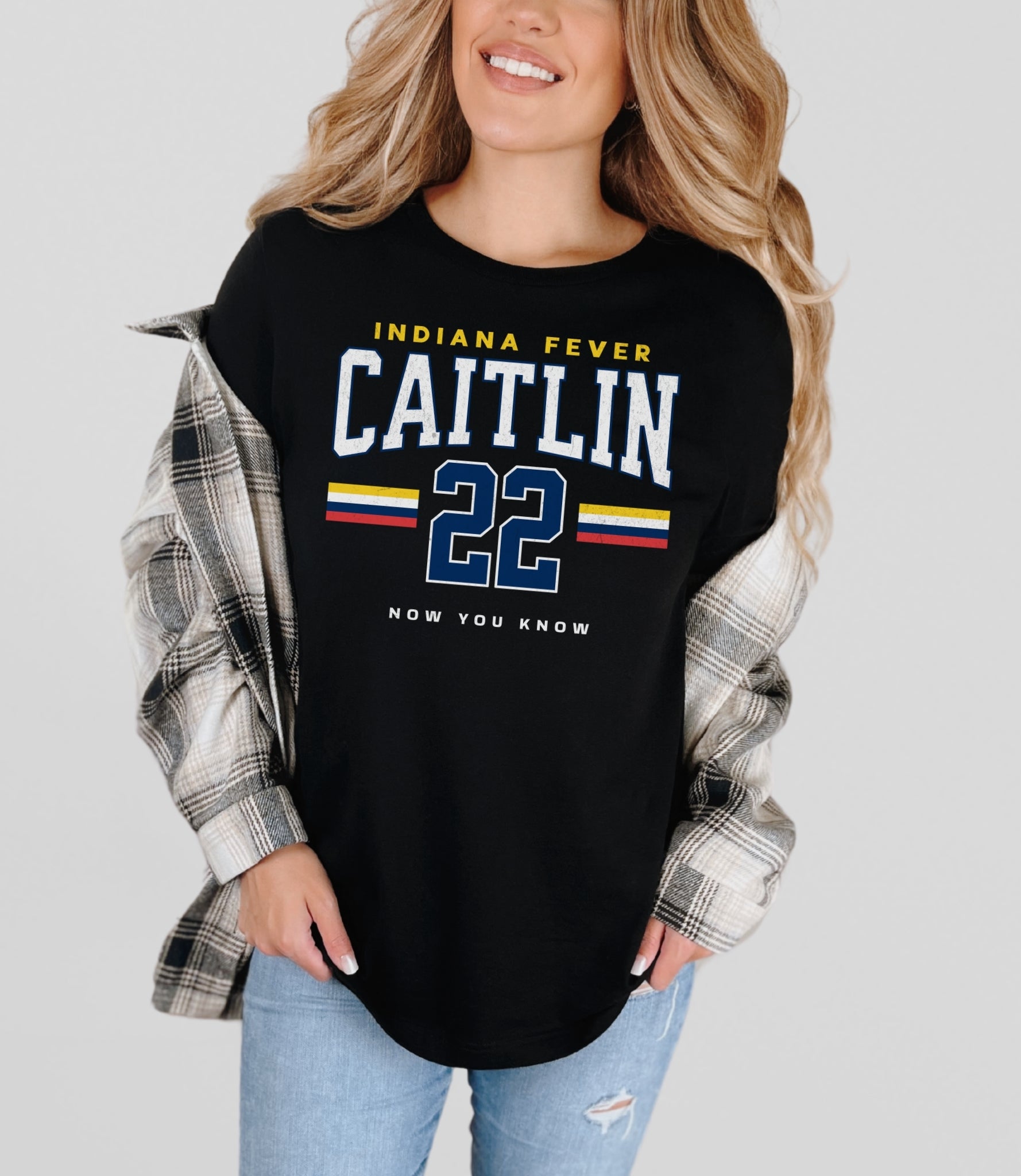 Woman wearing a black shirt with 'Indiana Fever Caitlin 22' text, flannel sleeves, and jeans on a light gray background.