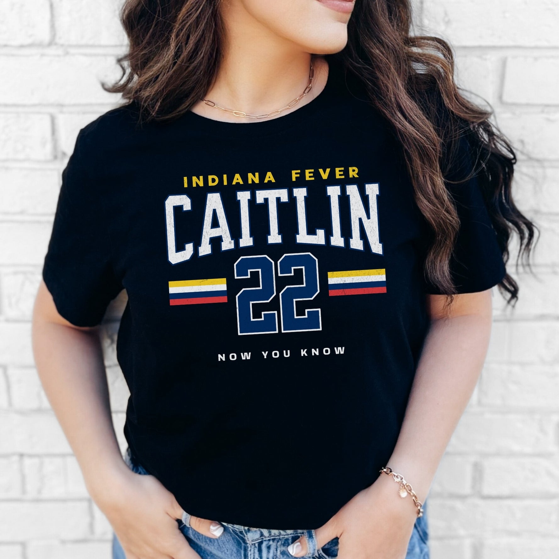 Person wearing a black t-shirt with 'Indiana Fever Caitlin 22 Now You Know' text against a white brick wall.