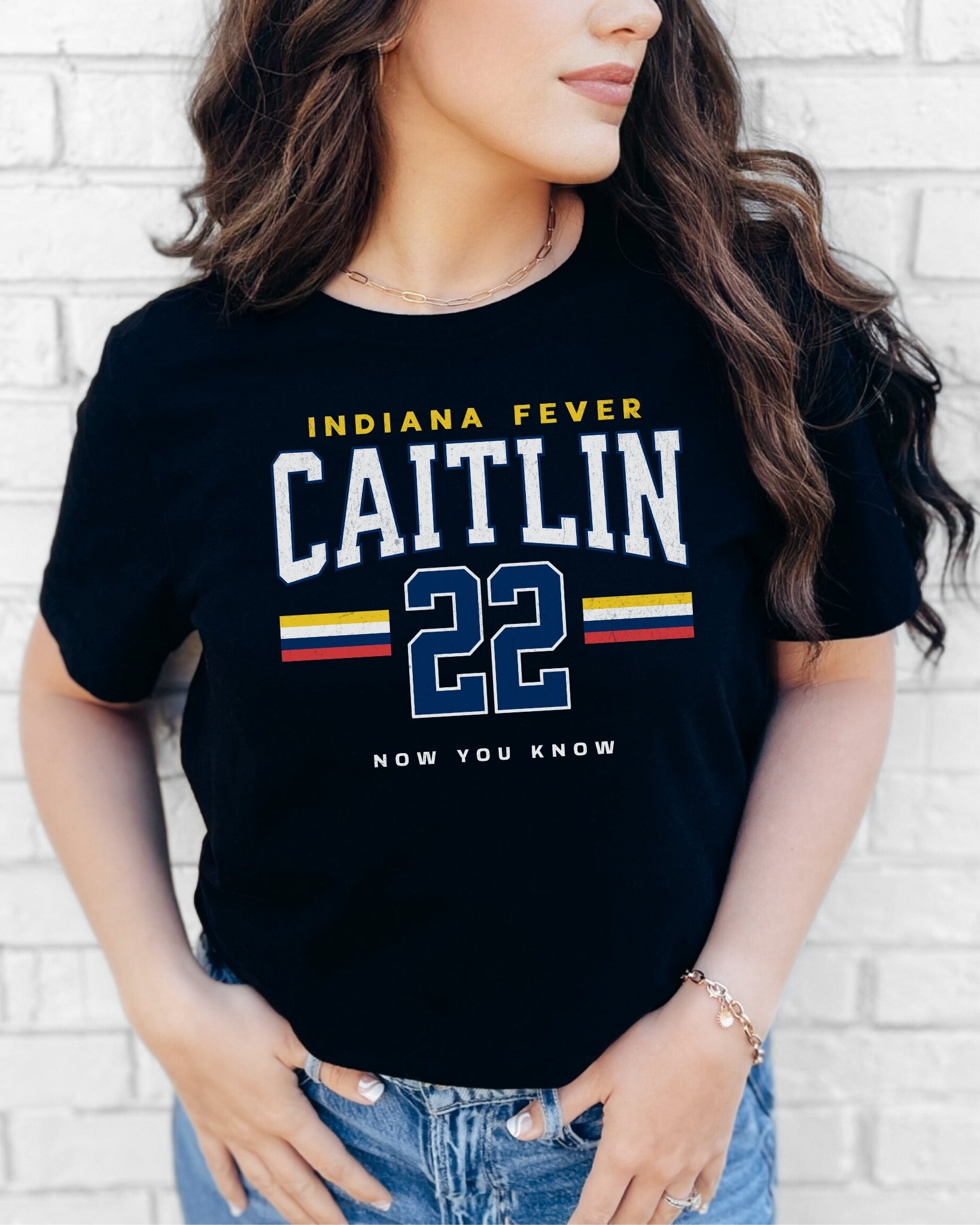 Person wearing a black t-shirt with 'Indiana Fever Caitlin 22 Now You Know' text against a white brick wall.