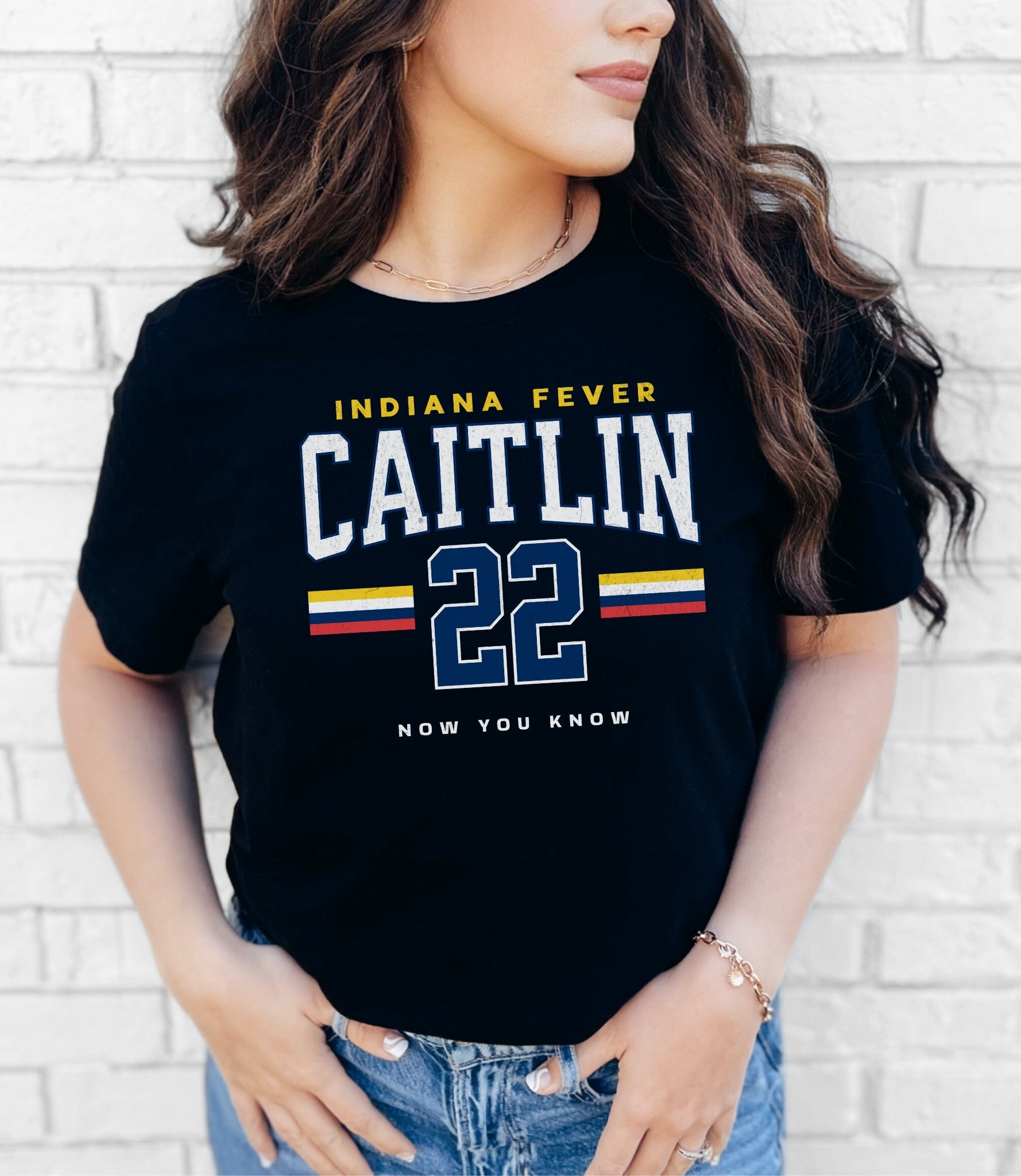 Person wearing a black t-shirt with 'Indiana Fever Caitlin 22 Now You Know' text against a white brick wall.
