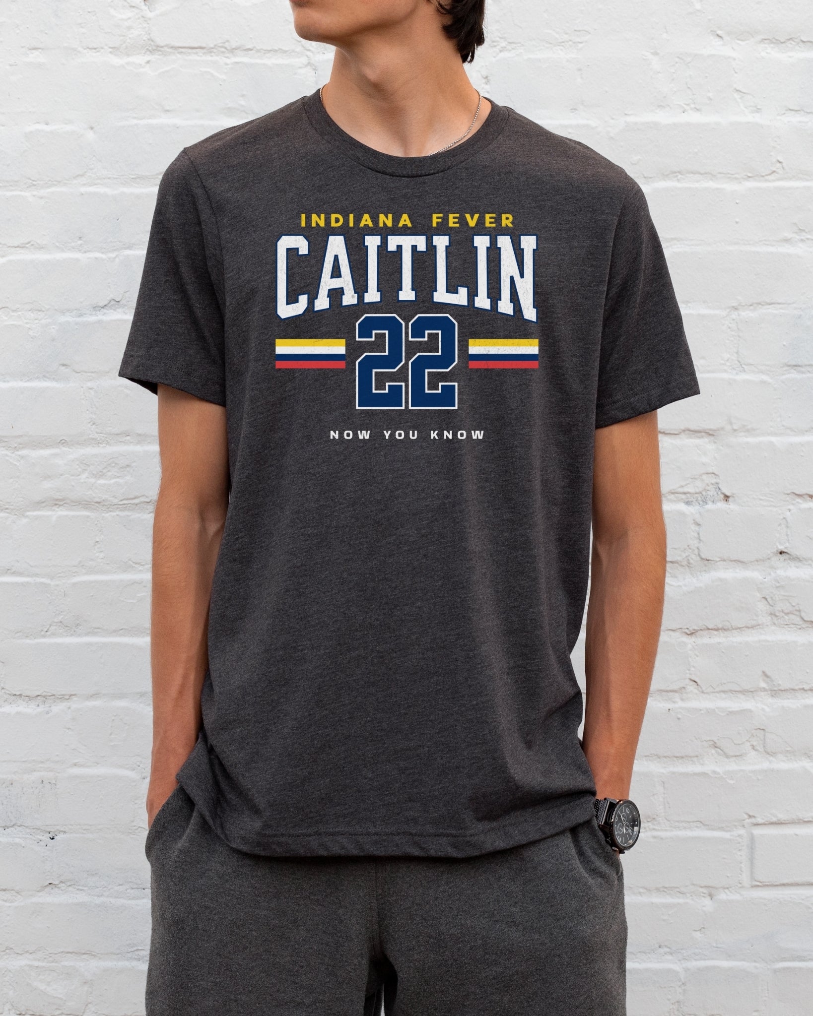 Person wearing a dark gray t-shirt with 'Indiana Fever Caitlin 22' text against a white brick wall.