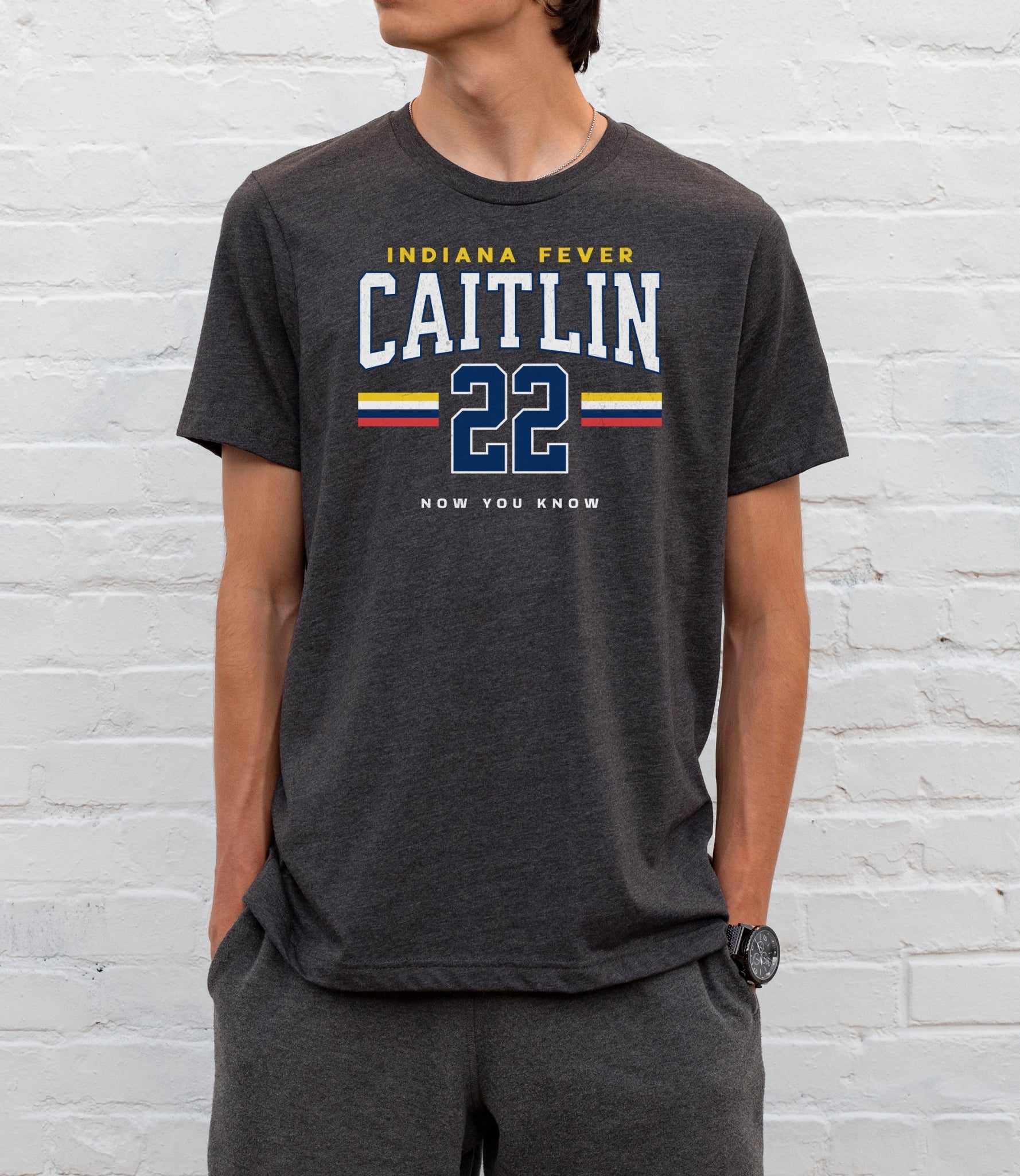 Person wearing a dark gray t-shirt with 'Indiana Fever Caitlin 22' text against a white brick wall.