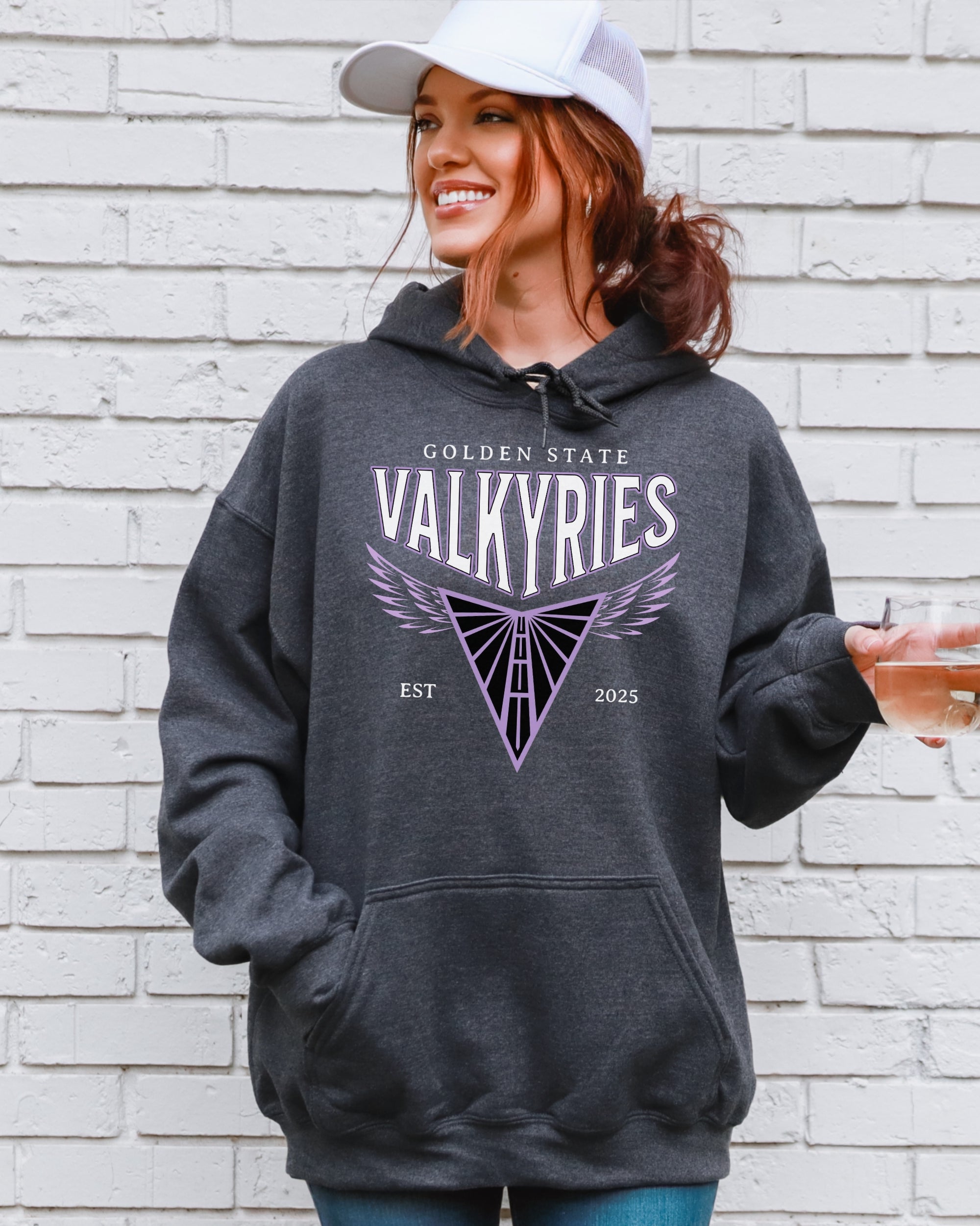 Woman wearing a gray hoodie with 'Golden State Valkyries' design against a white brick wall.
