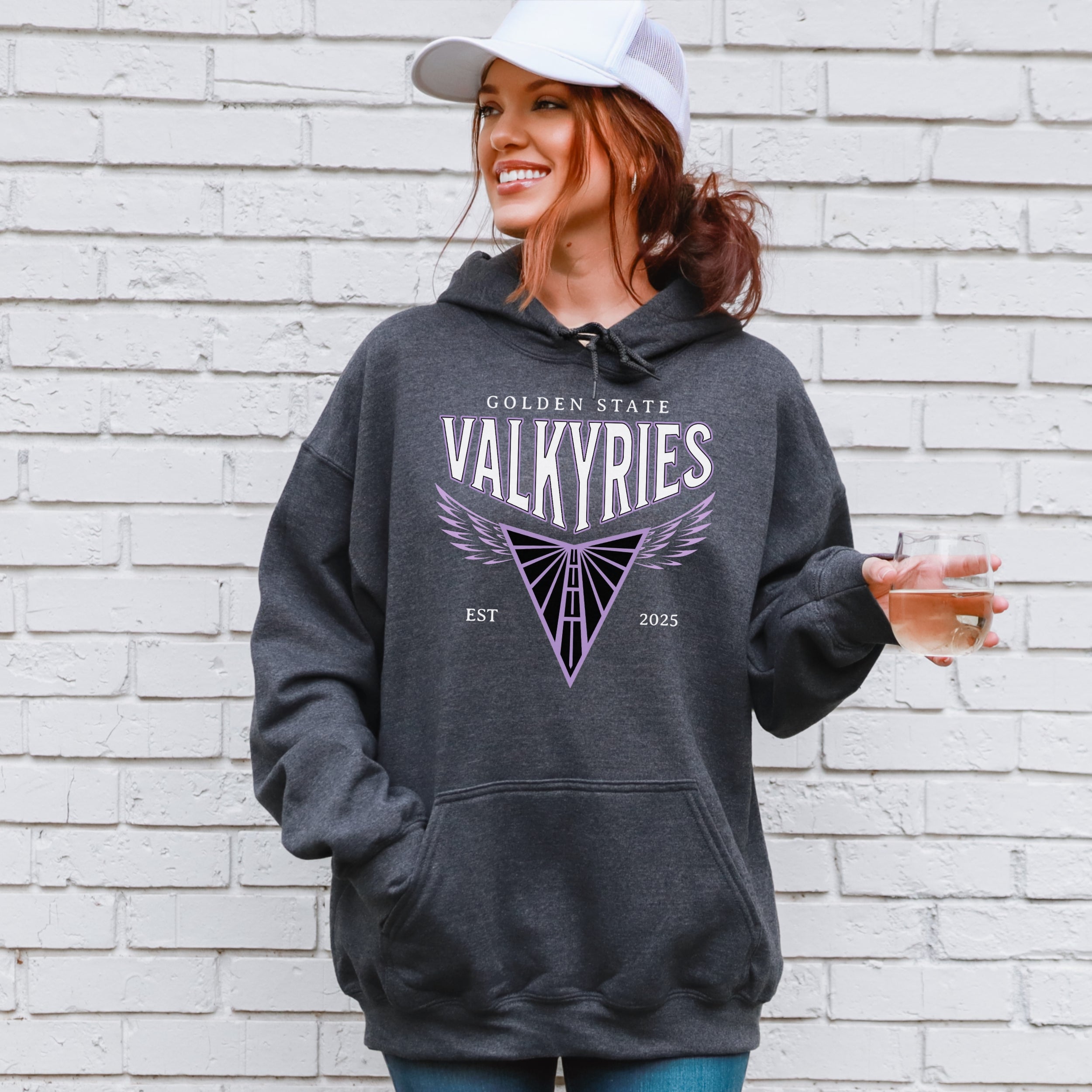 Woman wearing a gray hoodie with 'Golden State Valkyries' design against a white brick wall.