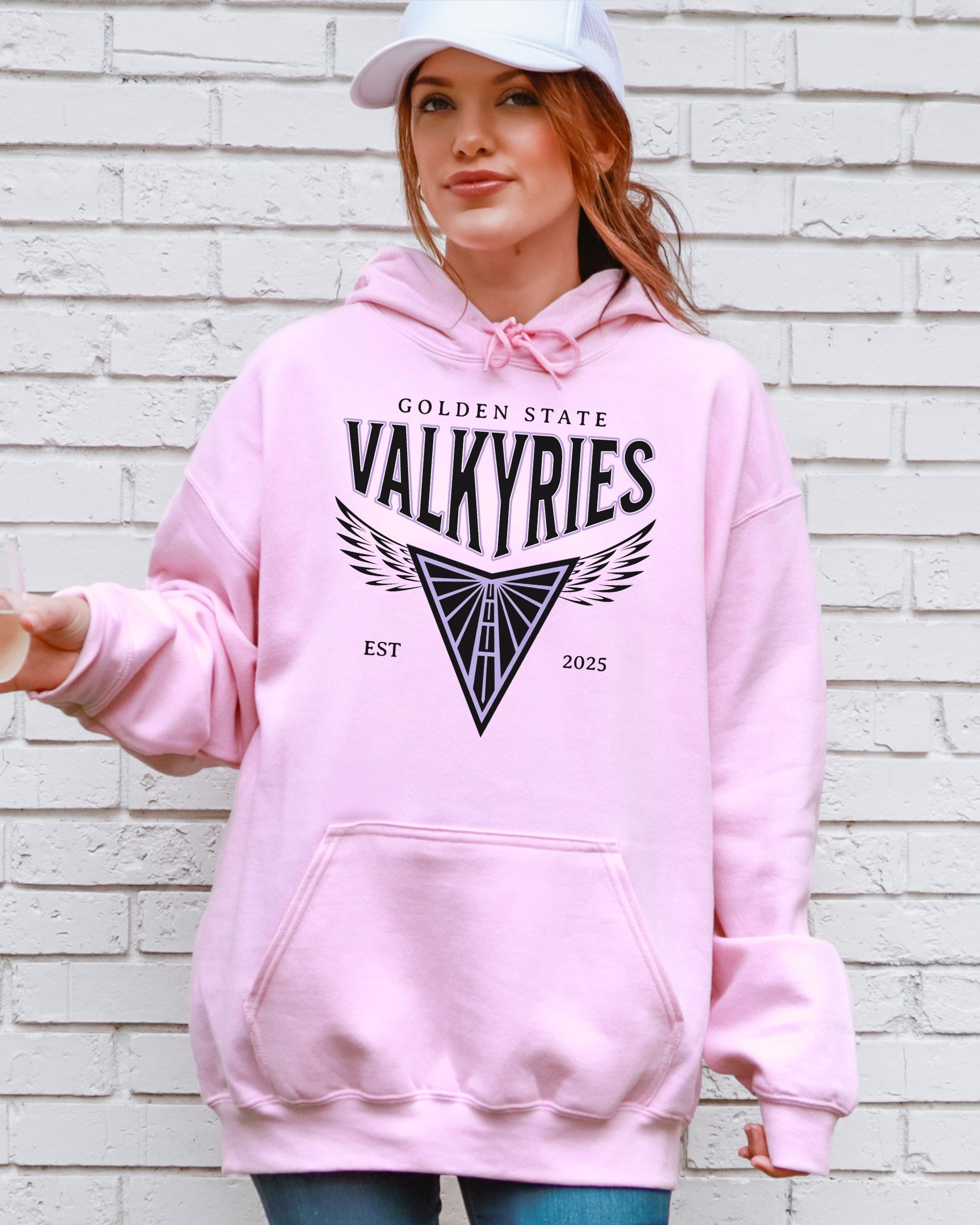 Person wearing a pink hoodie with 'Golden State Valkyries' design against a white brick wall.