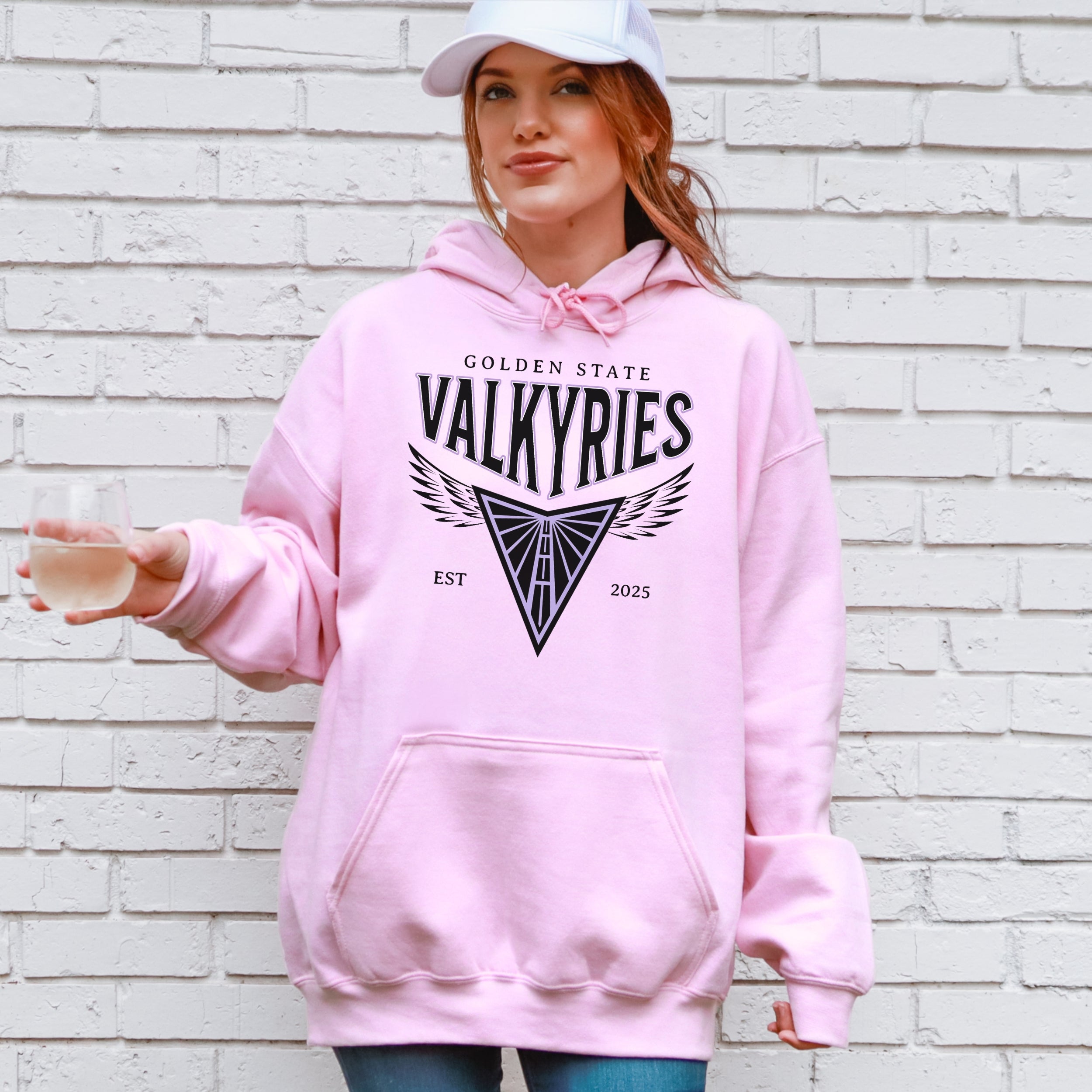 Person wearing a pink hoodie with 'Golden State Valkyries' design against a white brick wall.