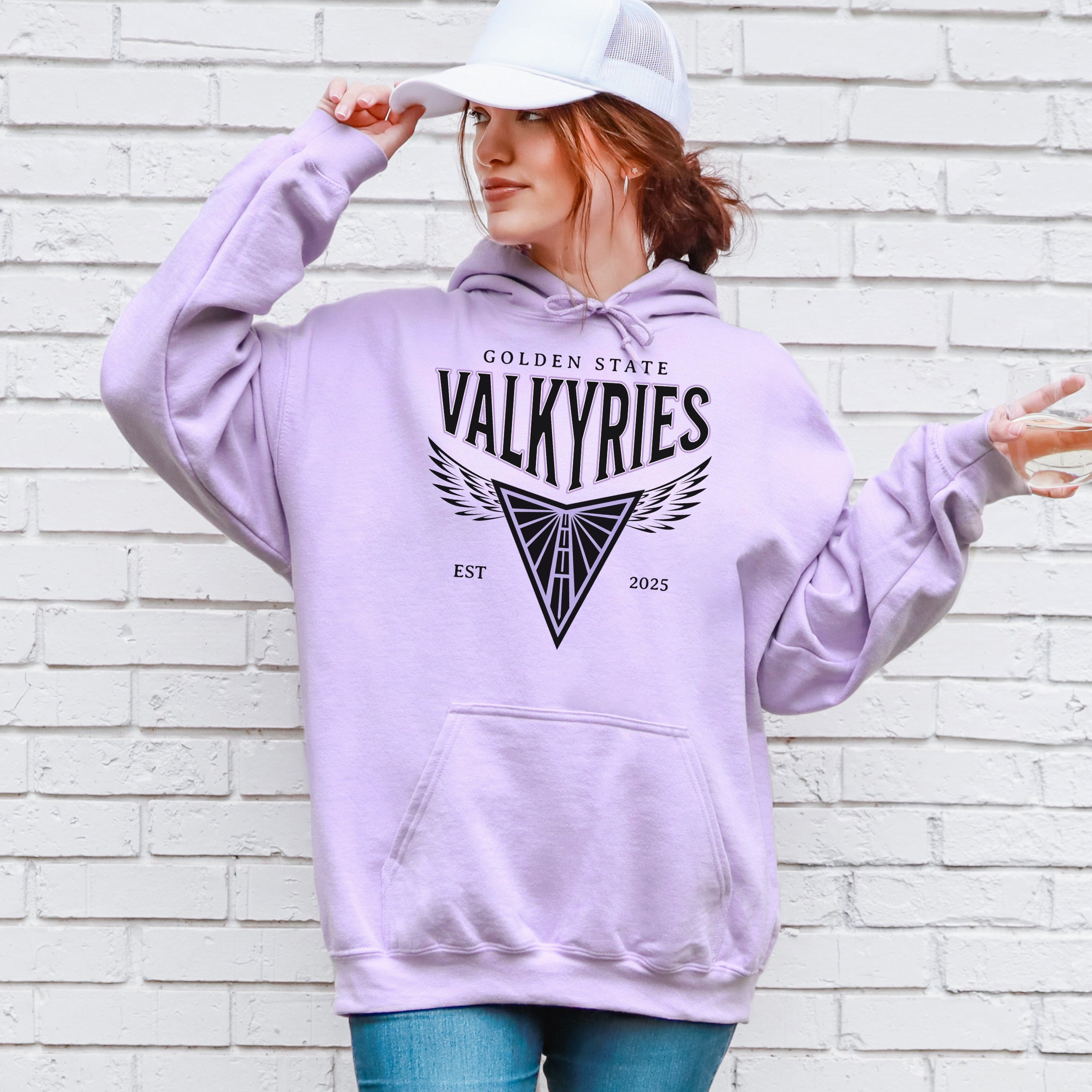 Person wearing a lavender hoodie with 'Golden State Valkyries' text and design, standing against a white brick wall.