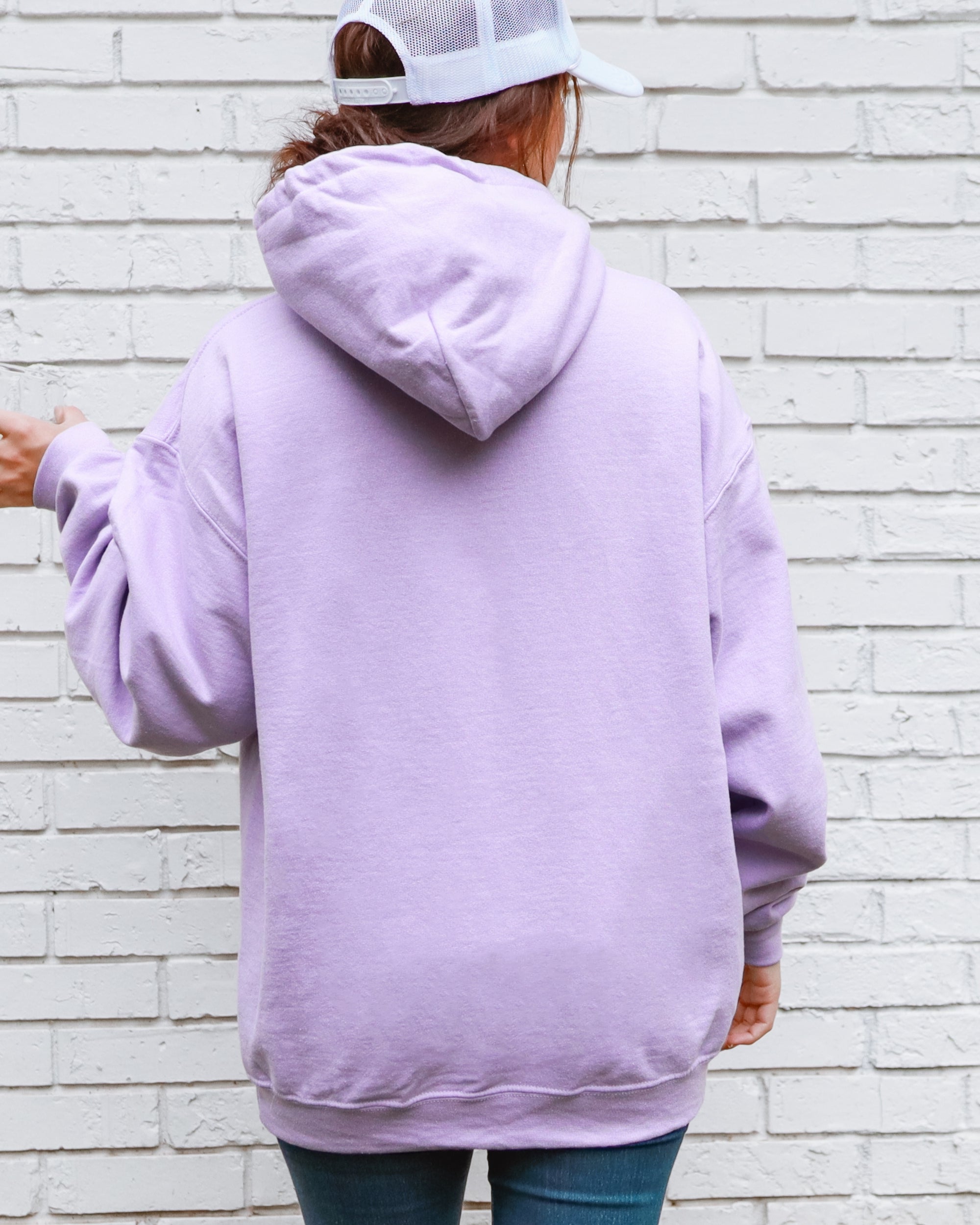 Person wearing a light purple hoodie and white cap, standing against a white brick wall.
