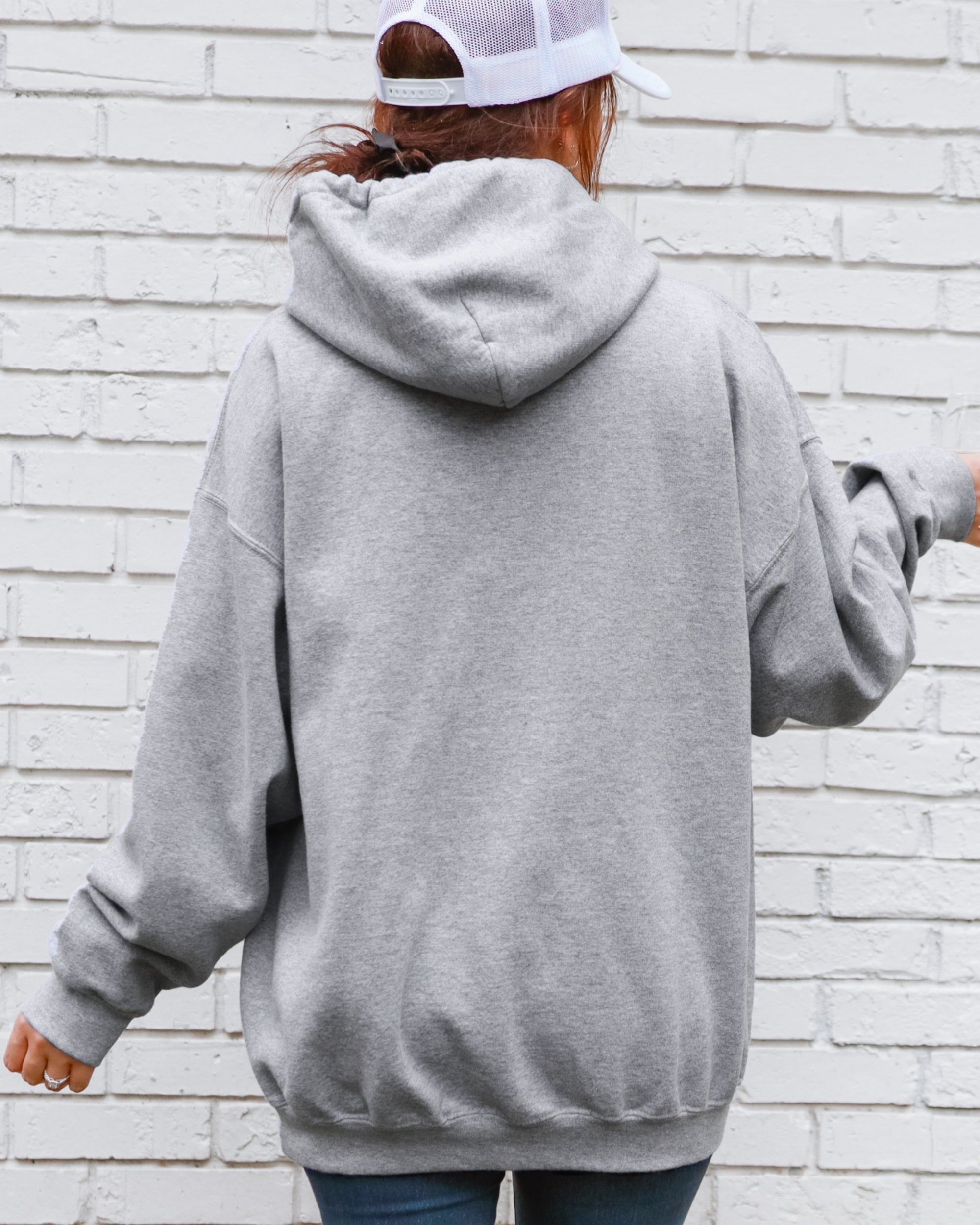 Person wearing a gray hoodie and white cap from behind against a light brick wall.