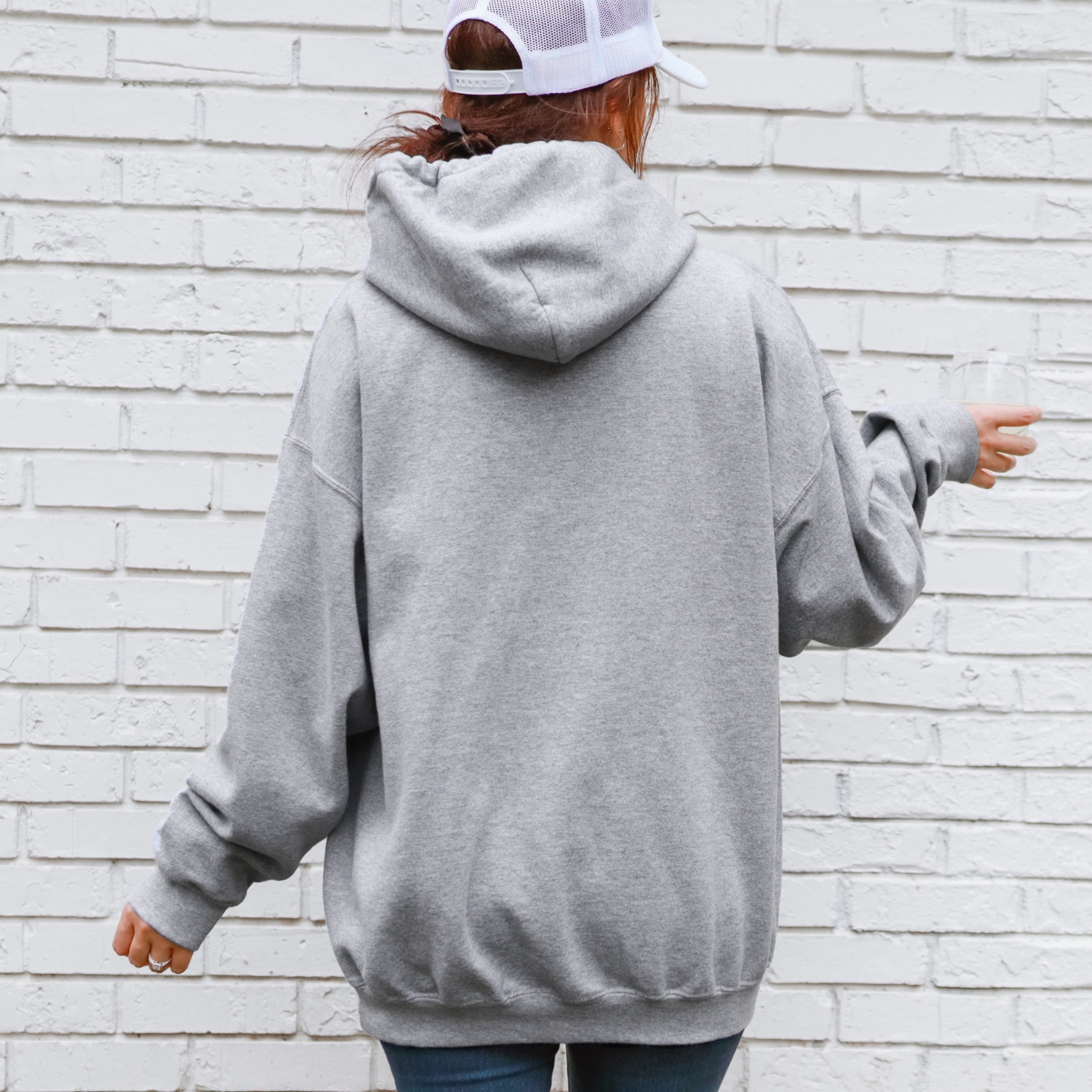 Person wearing a gray hoodie and white cap from behind against a light brick wall.