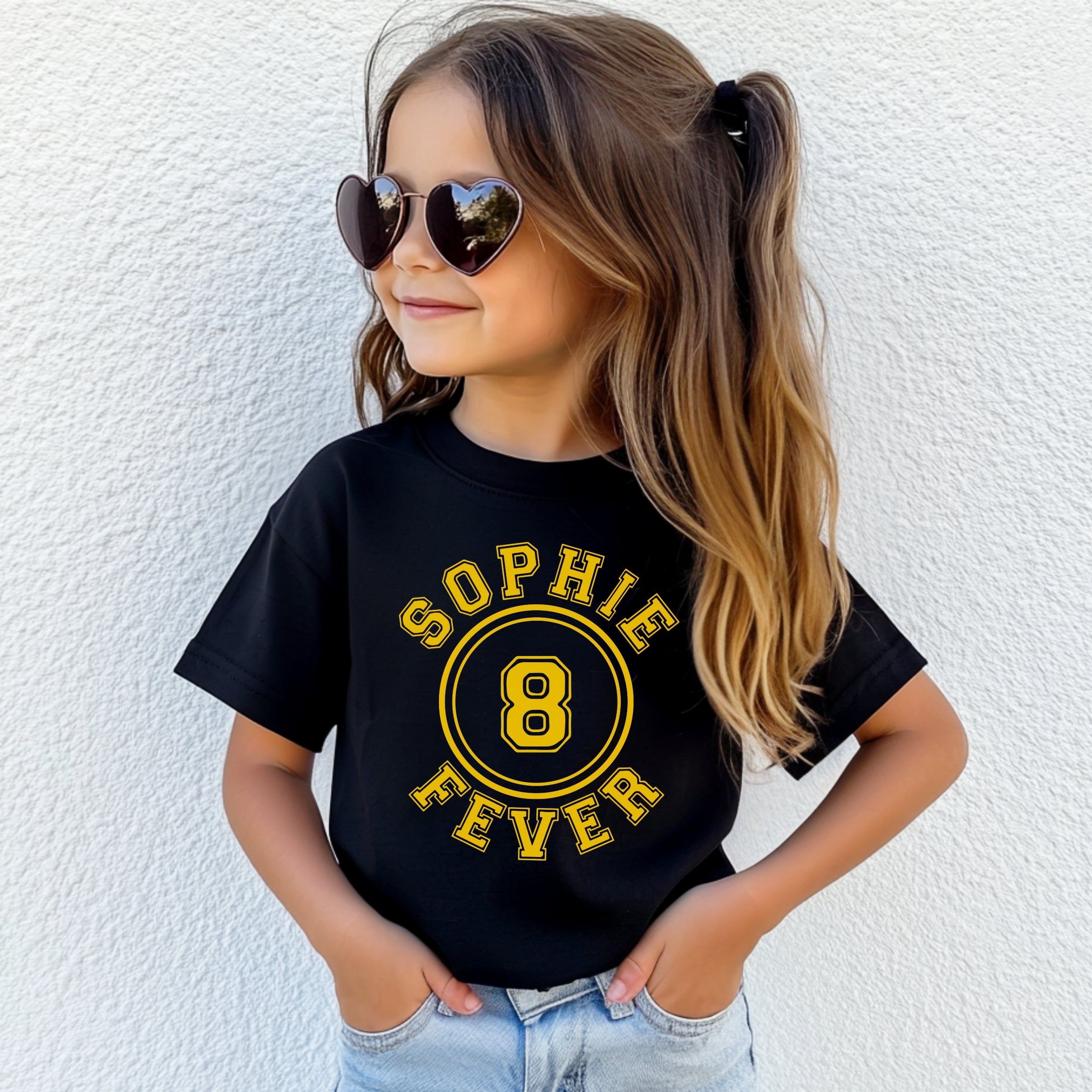 Child wearing a black t-shirt with 'Sophie Fever 8' in yellow, standing against a white wall.