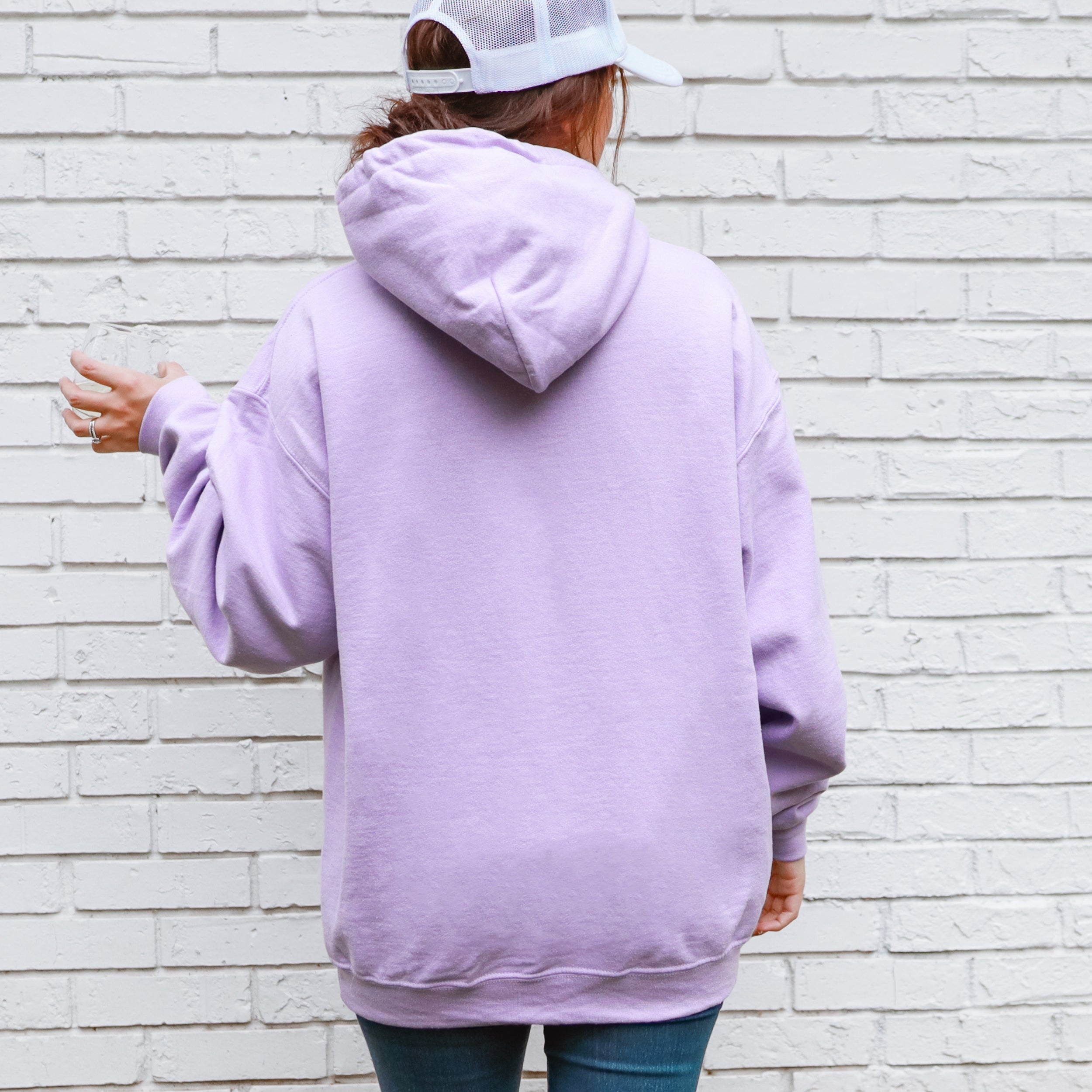 Person wearing a light purple hoodie and white cap, standing against a white brick wall.