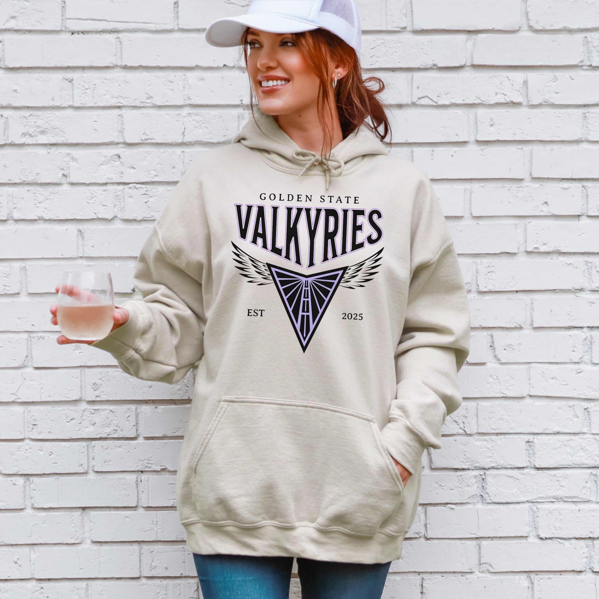 Person wearing a beige hoodie with 'Golden State Valkyries' design against a white brick wall.