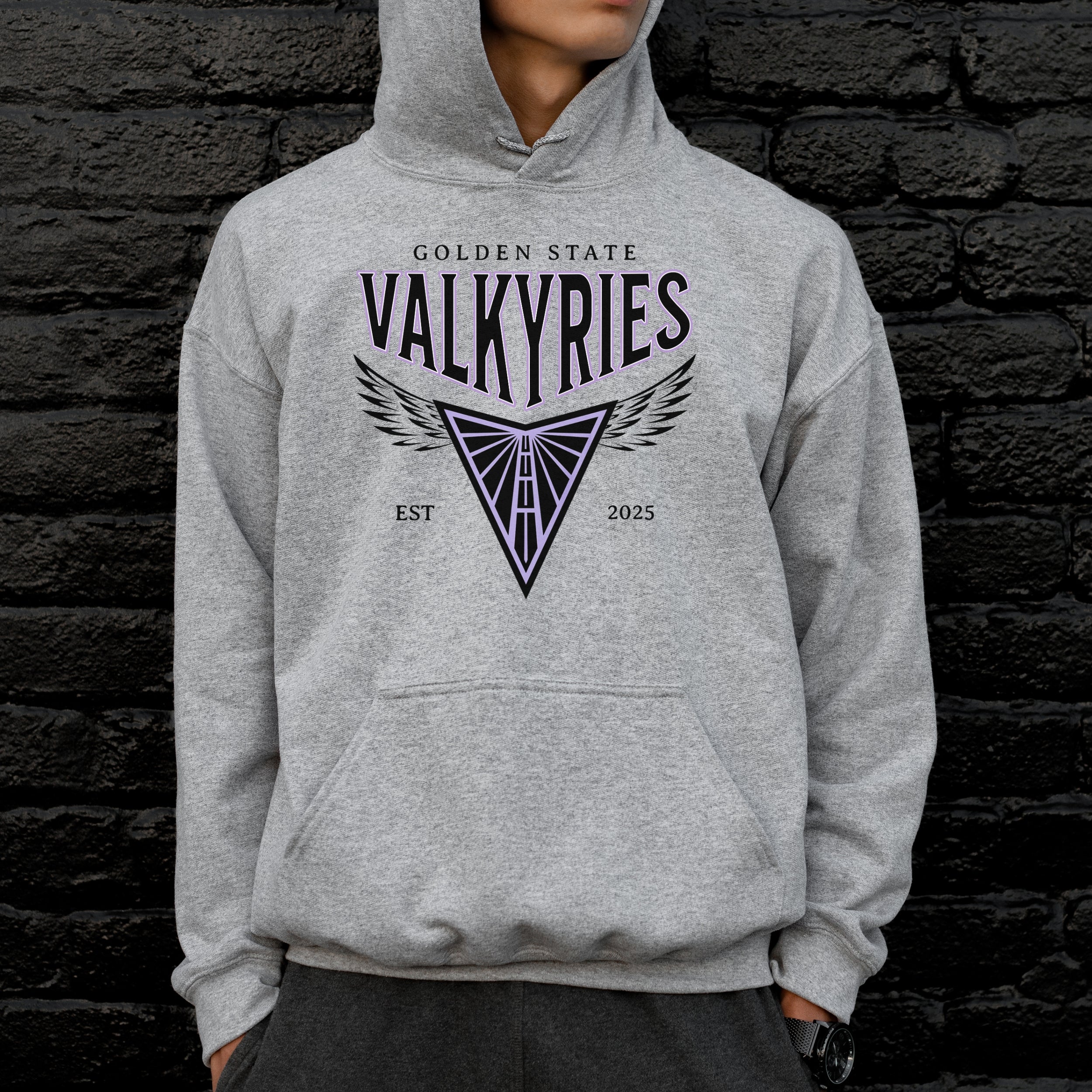 Person wearing a gray hoodie with 'Golden State Valkyries' design against a dark brick wall.